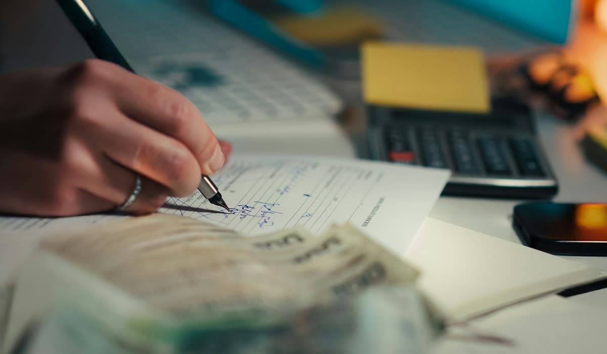A hand writing in a ledger beside cash and a calculator